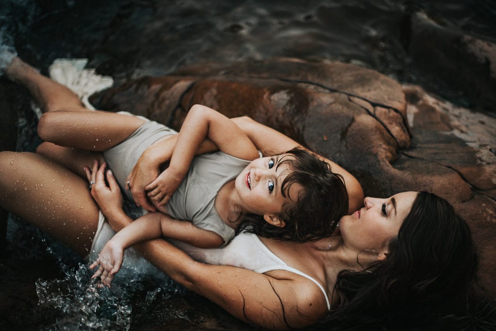 Online Family Photographer Education, A mother lays on rocks near the ocean with her baby girl who smiles in mom's care