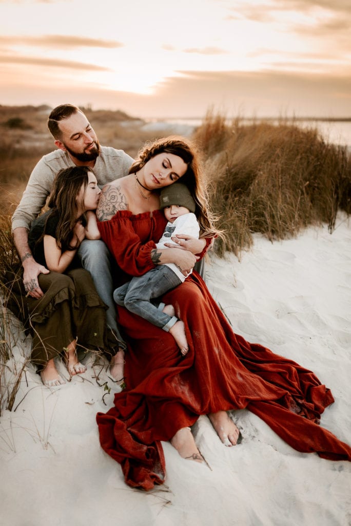 Online Family Photographer Education, A mother, father, daughter, and son snuggle together in the grass above the beach sand