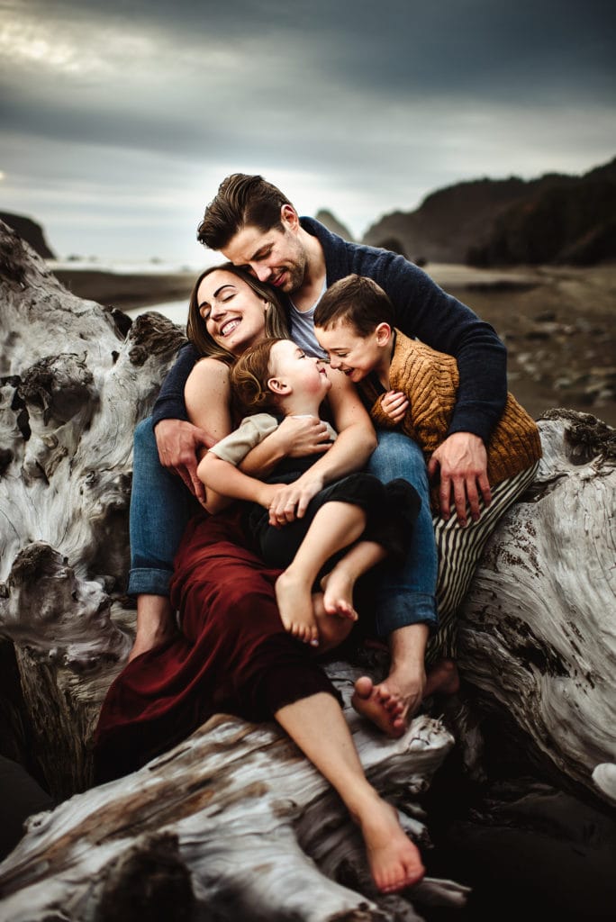 Online Family Photographer Education, A young family huddles together happy on rocks near the ocean
