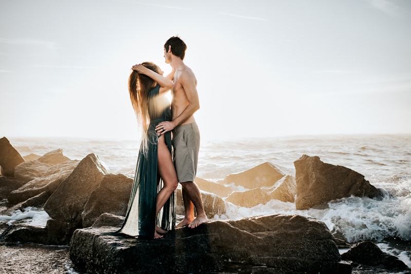 Online Family Photographer Education, a man and woman hold each other on ocean rocks as the waves crash around them