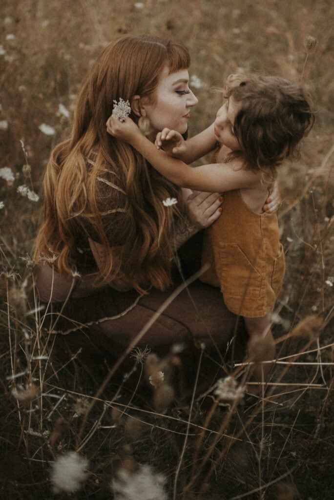 Mother kneeled down in tall grass with queen Anne's lace flowers next to her son. He's putting one of them behind her ear and has long red hair with curls. Motherhood moment from photo session in Boulder Colorado.